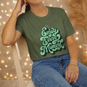 Peut inclure: T-shirt vert olive avec l'inscription "Enjoy Every Moment" dans une police rétro. Le texte est en nuances de vert avec un contour noir. Le t-shirt est porté avec un jean bleu.