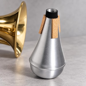 May include: A silver-colored metal trumpet mute with a conical shape, featuring a black felt ring at the top opening and three vertical cork strips attached to the upper section. A portion of a polished gold-colored brass trumpet bell is visible.