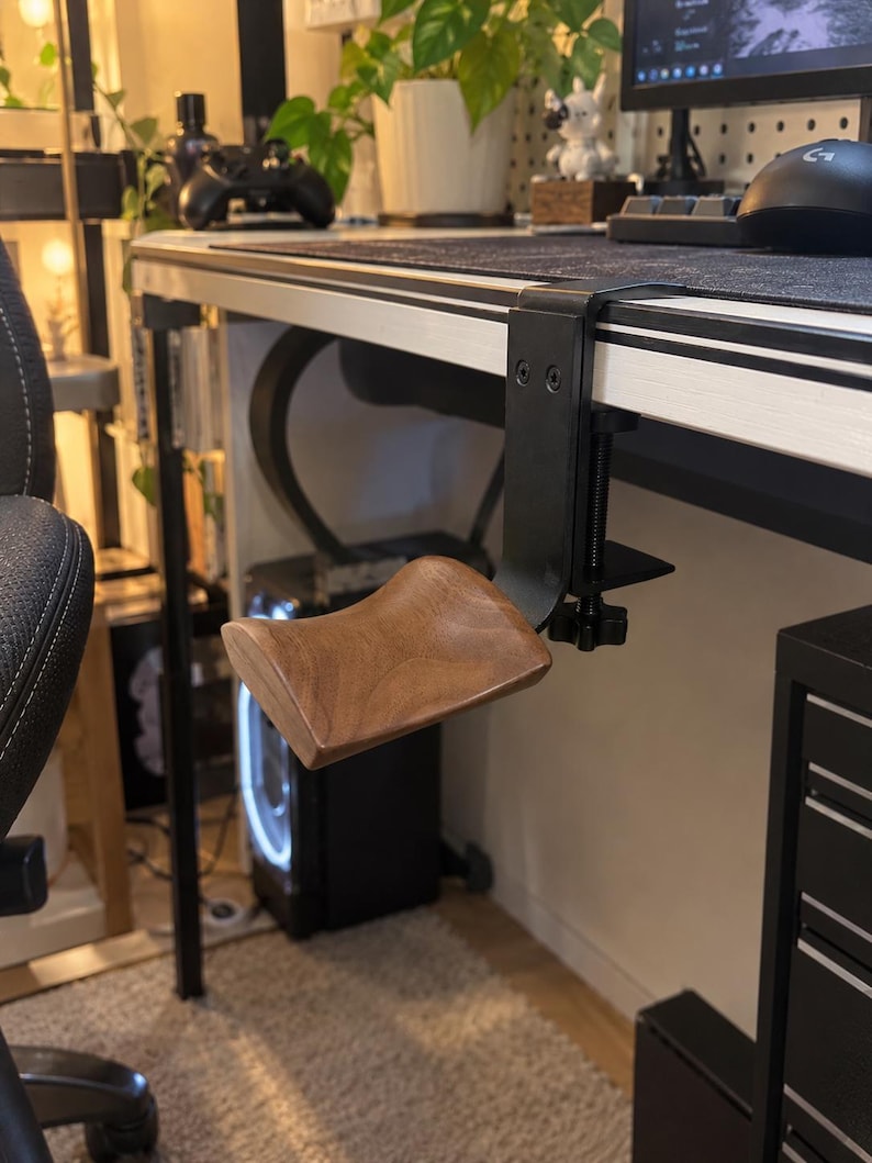 May include: A desk-mounted armrest featuring a contoured, dark brown wooden support pad attached to a black metal C-clamp mechanism. The clamp includes a vertical screw adjustment knob, designed to secure the unit to the edge of a flat desk surface.