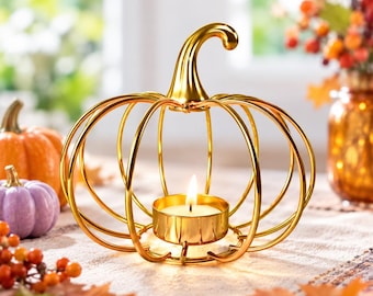 May include: A gold-coloured metal pumpkin-shaped candle holder with a wire frame design. The centre features a small circular cup holding a lit tea light candle. The structure is composed of curved metallic ribs forming a hollow pumpkin shape.