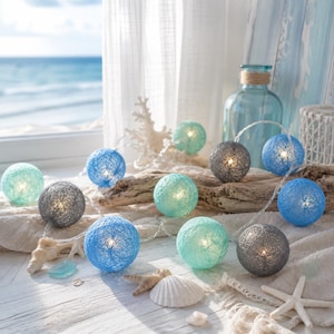 May include: A string of decorative globe lights in shades of blue, gray, and teal. The lights are arranged on a piece of driftwood, with seashells and a coastal background. The lights are lit, creating a warm glow.