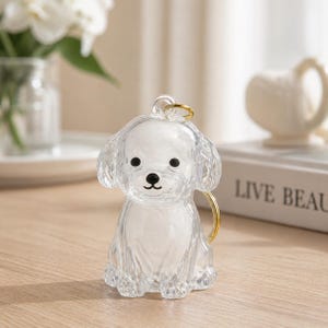 May include: A clear glass dog ornament with a gold-coloured ring attached to its head. The dog has a smiling face with black eyes and a black nose. The ornament is sitting on a wooden surface.