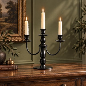 May include: A matte black metal candelabra featuring three arms with cylindrical candle cups. Three off-white taper candles are inserted into the cups, each topped with a lit flame. The candelabra has a central pedestal base and curved arms.