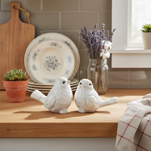 May include: Two white carved bird figurines sit on a wooden surface. Behind them are stacked plates with blue floral designs, a small potted succulent, and a jar of lavender. A wooden cutting board and a window are also visible.