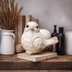 May include: A cream-coloured, carved wooden bird ornament, resting on a book bound with string. The bird has dark eyes and a textured finish. The background features decorative items such as books and vases.