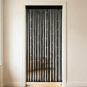 May include: Black beaded door curtain with white accents, hanging in a white door frame. The curtain is made of vertical strands of black beads, with small white beads interspersed throughout. The curtain hangs in front of a room with a wooden floor.