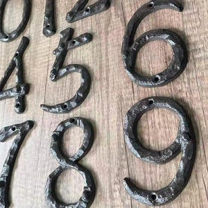 May include: Close-up of several black metal house numbers, including 0, 1, 4, 5, 6, 7, 8, and 9. The numbers have a rustic, textured appearance and are mounted on a light brown wooden surface. Each number has pre-drilled holes for easy installation.