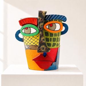 May include: A multi-coloured ceramic vase shaped like a stylised face with a central dark nose, red lips, and asymmetrical eyes. The surface features geometric patterns in green, yellow, blue, and orange, with a textured finish throughout.
