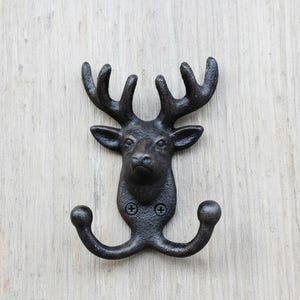 May include: A cast iron wall hook in the shape of a deer head. The hook is dark brown with two curved hooks below the deer's face. The deer has detailed antlers and is mounted on a light wood surface.