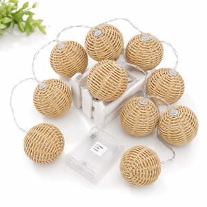 May include: A string of decorative lights featuring woven rattan spheres. Each sphere is a warm beige color and is illuminated by a small light. The lights are connected by a thin, silver wire and are powered by a battery pack.