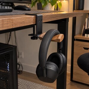 May include: A black metal desk clamp headphone stand featuring a natural wood grain horizontal arm. The clamp is secured to the edge of a wooden desk surface with a screw mechanism, holding a pair of black over-ear headphones by the headband.