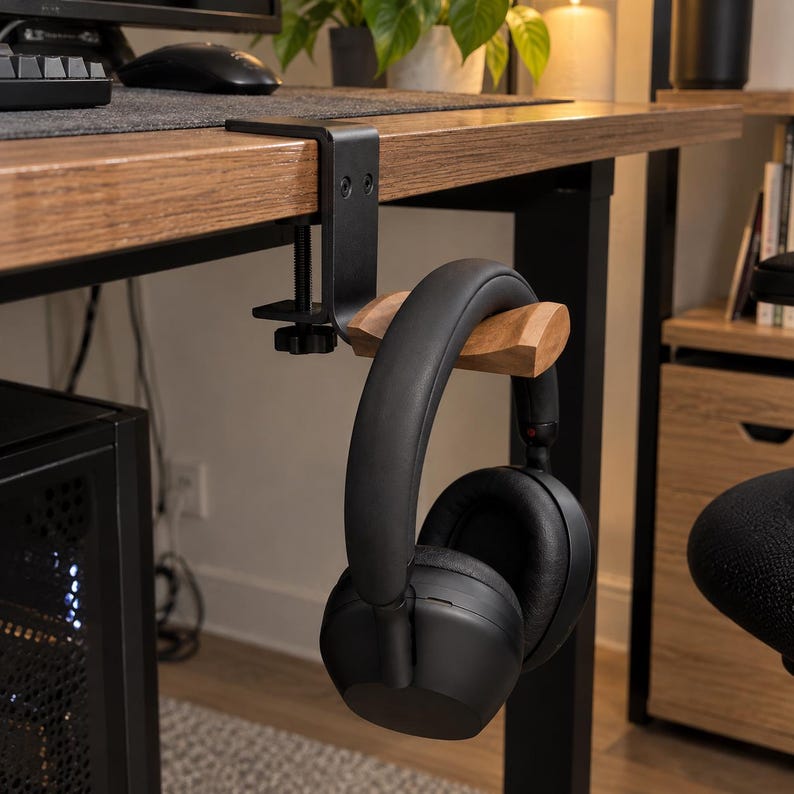May include: A black metal desk clamp headphone stand featuring a natural wood grain horizontal arm. The clamp is secured to the edge of a wooden desk surface with a screw mechanism, holding a pair of black over-ear headphones by the headband.