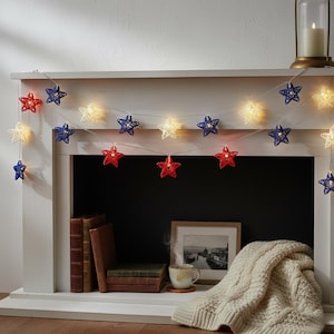 May include: A string of star-shaped fairy lights in red, white, and blue, draped across a white mantel. The lights are illuminated, creating a warm glow. The mantel also features books, a framed picture, and a candle.