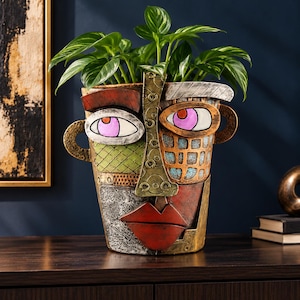 Puede incluir: Una maceta decorativa con forma de rostro humano y acabado texturizado multicolor. El rostro presenta ojos grandes con pupilas rosas, una nariz prominente y labios rojos. Una planta verde crece desde la parte superior del recipiente.