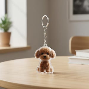 Puede incluir: Un llavero acrílico transparente con un diseño de perro caniche marrón. El perro tiene pelaje esponjoso y una cara sonriente. El llavero tiene una cadena y un anillo plateados. El perro está sentado en una mesa de madera.