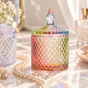 Puede incluir: Un frasco cilíndrico de vidrio con un patrón de corte de diamante y un acabado iridiscente que cambia entre tonos rosas, amarillos y verdes. El frasco cuenta con una tapa extraíble a juego con un remate puntiagudo y facetado.