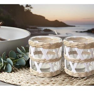 May include: Two white wicker candle holders with a woven design. The candle holders are on a woven mat and are in front of a blurry background of a beach and ocean.