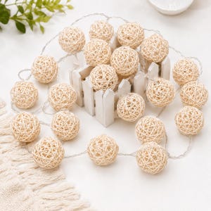 May include: String lights with woven rattan spheres. The spheres are a light beige colour and are strung on a thin, silver wire. The lights are arranged on a white surface with a small white picket fence.