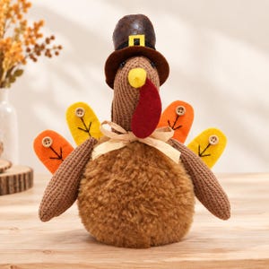 May include: A plush turkey figurine featuring a brown textured body, a dark brown hat with a yellow buckle, and a red wattle. The tail consists of four felt feathers in yellow and orange, each accented with a small wooden button and a stitched leaf.