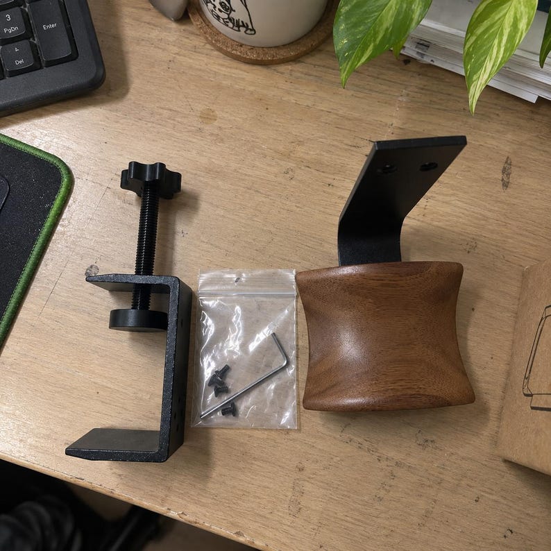 May include: A black metal C-clamp with a threaded adjustment knob, a small clear plastic bag containing four black screws and an L-shaped hex key, and a curved wooden headphone stand mount with a black metal bracket attached to the top.
