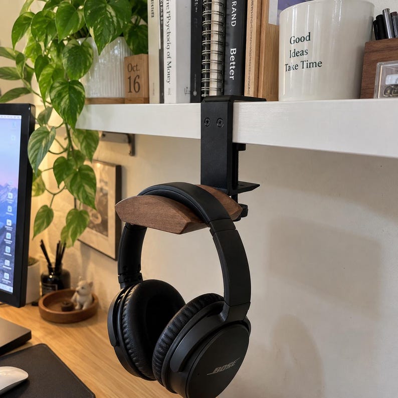 May include: A black metal headphone stand with a dark wood curved rest, featuring a clamp mechanism attached to a white shelf. The stand holds a pair of black over-ear headphones with cushioned ear cups and a branded headband.
