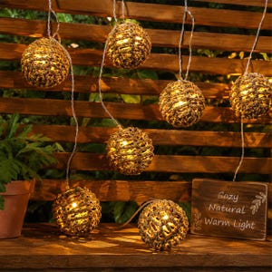 May include: String lights with woven, spherical shades, illuminated with warm white lights. The lights hang against a wooden fence and a wooden sign that reads "Cozy Natural Warm Light". The lights are a decorative element for outdoor spaces.
