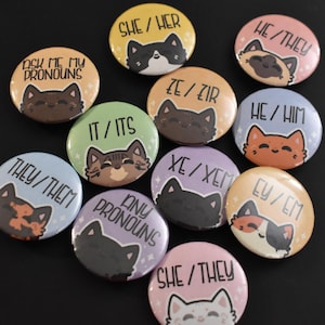 May include: A selection of pronoun badges with cat designs. Each badge shows a different pronoun set, including "She/Her," "He/Him," and "They/Them," alongside a cute cat illustration. The badges are arranged on a black background.