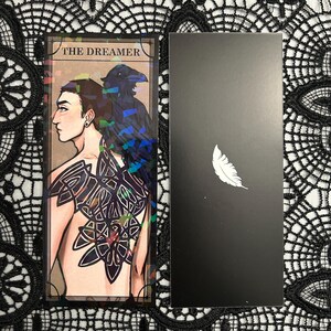 Op de afbeelding: Een tarotkaart met een illustratie van een persoon met een raaf en een ingewikkelde rugtatoeage, getiteld "THE DREAMER". Een zwarte kaart met een witte veer staat ernaast. De kaarten hebben een holografisch effect.