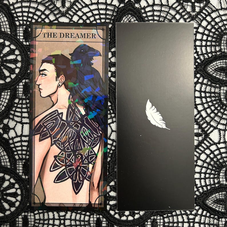 Op de afbeelding: Een tarotkaart met een illustratie van een persoon met een raaf en een ingewikkelde rugtatoeage, getiteld "THE DREAMER". Een zwarte kaart met een witte veer staat ernaast. De kaarten hebben een holografisch effect.