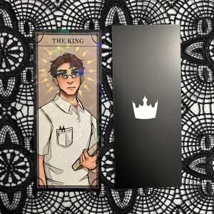 Op de afbeelding: Een tarotkaart met de tekst "THE KING" en een illustratie van een persoon met een bril en een wit overhemd, die een boek vasthoudt. Daarnaast een zwarte kaart met een witte kroon.
