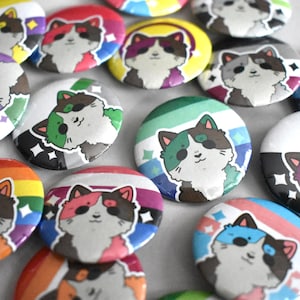 May include: A collection of colorful round buttons featuring a cartoon cat face. Each button has a different background design with various colors, including rainbow, pink, blue, and purple. The cat has a black and brown color scheme.
