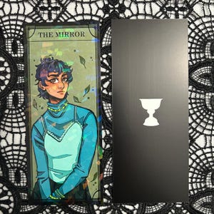 Op de afbeelding: Twee tarotkaarten. E&eacute;n kaart toont een illustratie van een persoon in teal kleding met de tekst "THE MIRROR". De andere kaart is zwart met een wit kelksymbool. De kaarten liggen op een zwart kanten oppervlak.