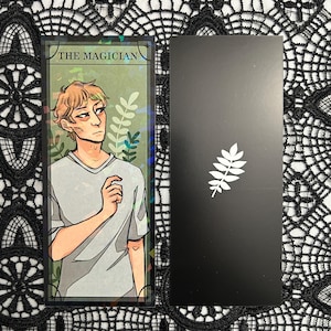Op de afbeelding: Twee tarotkaarten. E&eacute;n kaart toont een illustratie van een persoon met de tekst "THE MAGICIAN". De andere kaart is zwart met een wit bladeren ontwerp. De kaarten liggen op een zwart-wit kanten oppervlak.