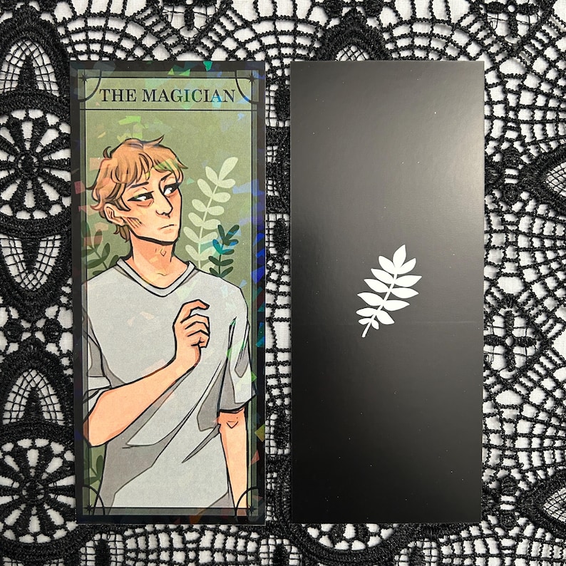 Op de afbeelding: Twee tarotkaarten. E&eacute;n kaart toont een illustratie van een persoon met de tekst "THE MAGICIAN". De andere kaart is zwart met een wit bladeren ontwerp. De kaarten liggen op een zwart-wit kanten oppervlak.