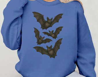 May include: A blue sweatshirt with a design of four bats in flight. The bats are illustrated in a vintage style, with detailed shading and a textured appearance. The bats are positioned to suggest movement, creating a dynamic composition.