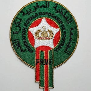 Op de afbeelding: Een geborduurde patch in groen en goud met een rode en witte ster en een gouden kroon in het midden. De tekst rond de ster luidt "Fédération Royale Marocaine de Football" in het Arabisch. De tekst "FRMF" staat onder de ster.