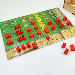 May include: A board game with a green game board featuring a map design, red game pieces, and a wooden storage box. The game pieces are various shapes, including cylinders, triangles, and rectangular blocks. The game board has a detailed design.