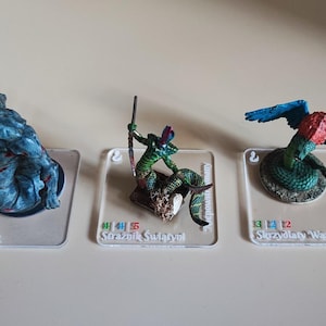 Pode incluir: Três figuras de fantasia em miniatura sobre bases transparentes. As figuras incluem um monstro azul, um humanoide verde com uma lança e uma serpente alada. Cada figura é pintada com cores e texturas detalhadas.