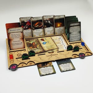 Könnte beinhalten: Ein hellbrauner, lasergeschnittener Brettspiel-Organizer mit Schlitzen für mehrere rechteckige Spielkarten, runde Spielsteine und kleine farbige Würfel. Der Organizer trägt die Gravur LuckyGRAW und enthält verschiedene Spielkomponenten, darunter Karten mit polnischem Text und kleine Kunststoffmarker.