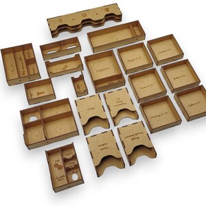 May include: A collection of laser-cut wooden storage components, including trays, boxes, and holders. Some pieces have text, such as "Phantas & Co." and "French Foreign Legion". The pieces are arranged on a white surface.