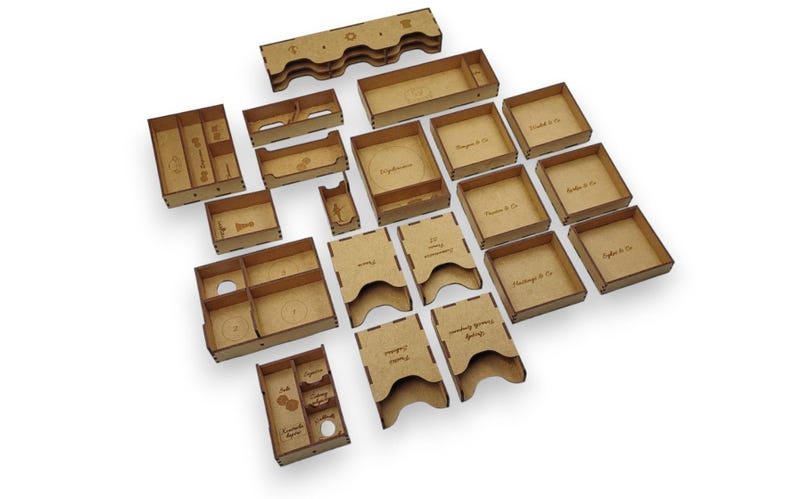 May include: A collection of laser-cut wooden storage components, including trays, boxes, and holders. Some pieces have text, such as "Phantas & Co." and "French Foreign Legion". The pieces are arranged on a white surface.