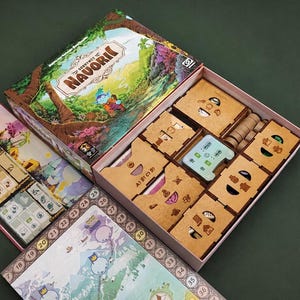 Può includere: Un gioco da tavolo intitolato Convergence of Navorii con un coperchio colorato raffigurante una scena boschiva. La scatola aperta contiene organizzatori in legno per i componenti, insieme a un tabellone con un percorso numerato e mappe.