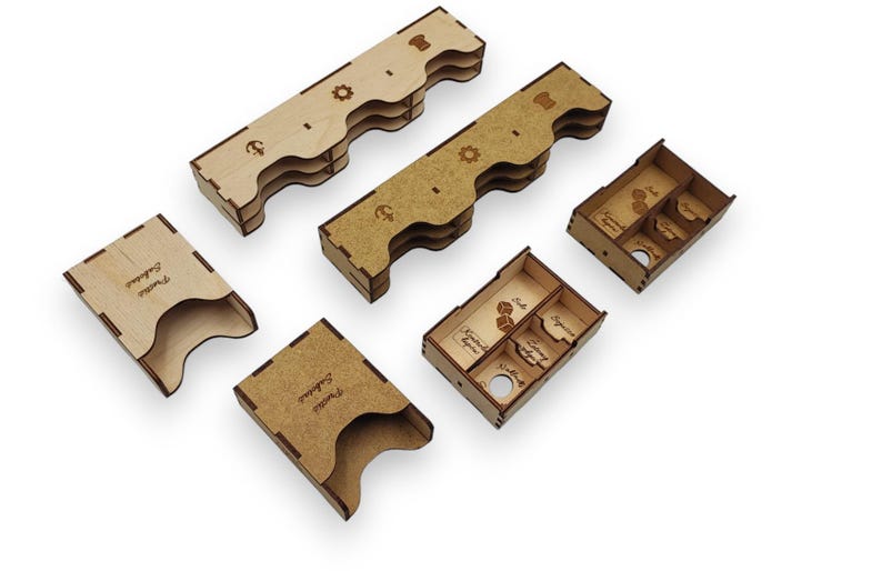 May include: A collection of laser-cut wooden game organizers. The set includes several rectangular trays, some with divided compartments, and two card holders. The organizers are light brown with engraved details and text.