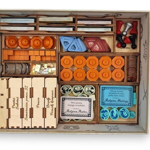 May include: Wooden game organizer with multiple compartments filled with game pieces. The pieces include orange tokens, blue cards, and wooden blocks. The organizer has text on the side and is designed to hold board game components.