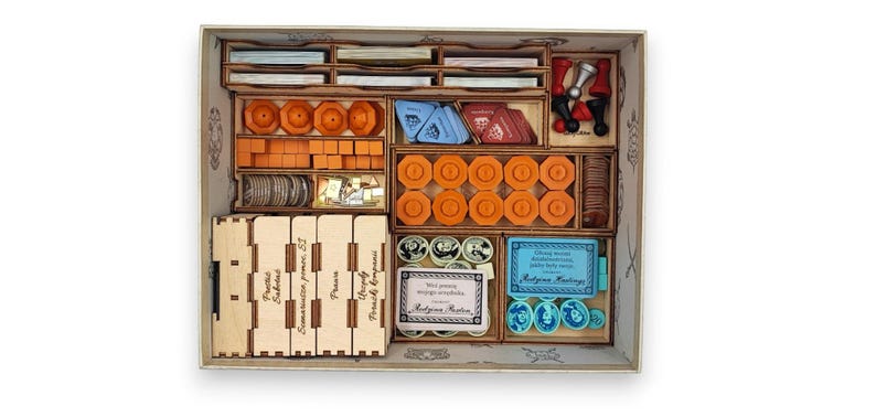 May include: Wooden game organizer with multiple compartments filled with game pieces. The pieces include orange tokens, blue cards, and wooden blocks. The organizer has text on the side and is designed to hold board game components.