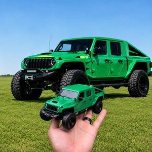 May include: A bright green four-door pickup truck with a black grille, large off-road tires, and a front bumper winch. A smaller, matching green scale model of the same pickup truck is held in the foreground, mirroring the design of the full-sized vehicle.