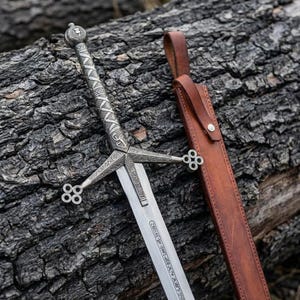 May include: A silver-toned metal sword featuring an intricate cross-guard with four circular accents at the ends and a textured handle. The blade displays engraved lettering near the hilt. A brown leather scabbard with a metal rivet is positioned nearby.