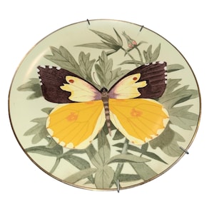 May include: A decorative plate featuring a detailed illustration of a butterfly with yellow and brown wings, set against a backdrop of green leaves and a light green background. The plate has a gold rim and is mounted with metal hangers.