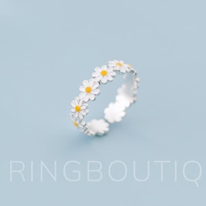 Può includere: Un anello d'argento con un motivo a fiori di margherita. L'anello ha petali bianchi e centri gialli. Il testo "RINGBOUTIQ" è nella parte inferiore dell'immagine. L'anello è posto su uno sfondo azzurro.
