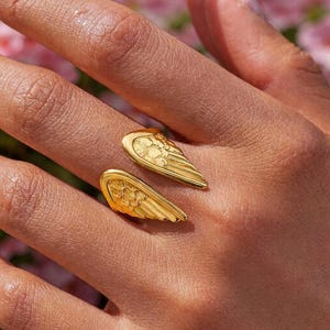 Op de afbeelding: Een goudkleurige ring met een vleugelontwerp. De ring heeft twee gedetailleerde vleugels die om de vinger wikkelen. De vleugels hebben een textuur. De achtergrond is een vervaging van roze bloemen.
